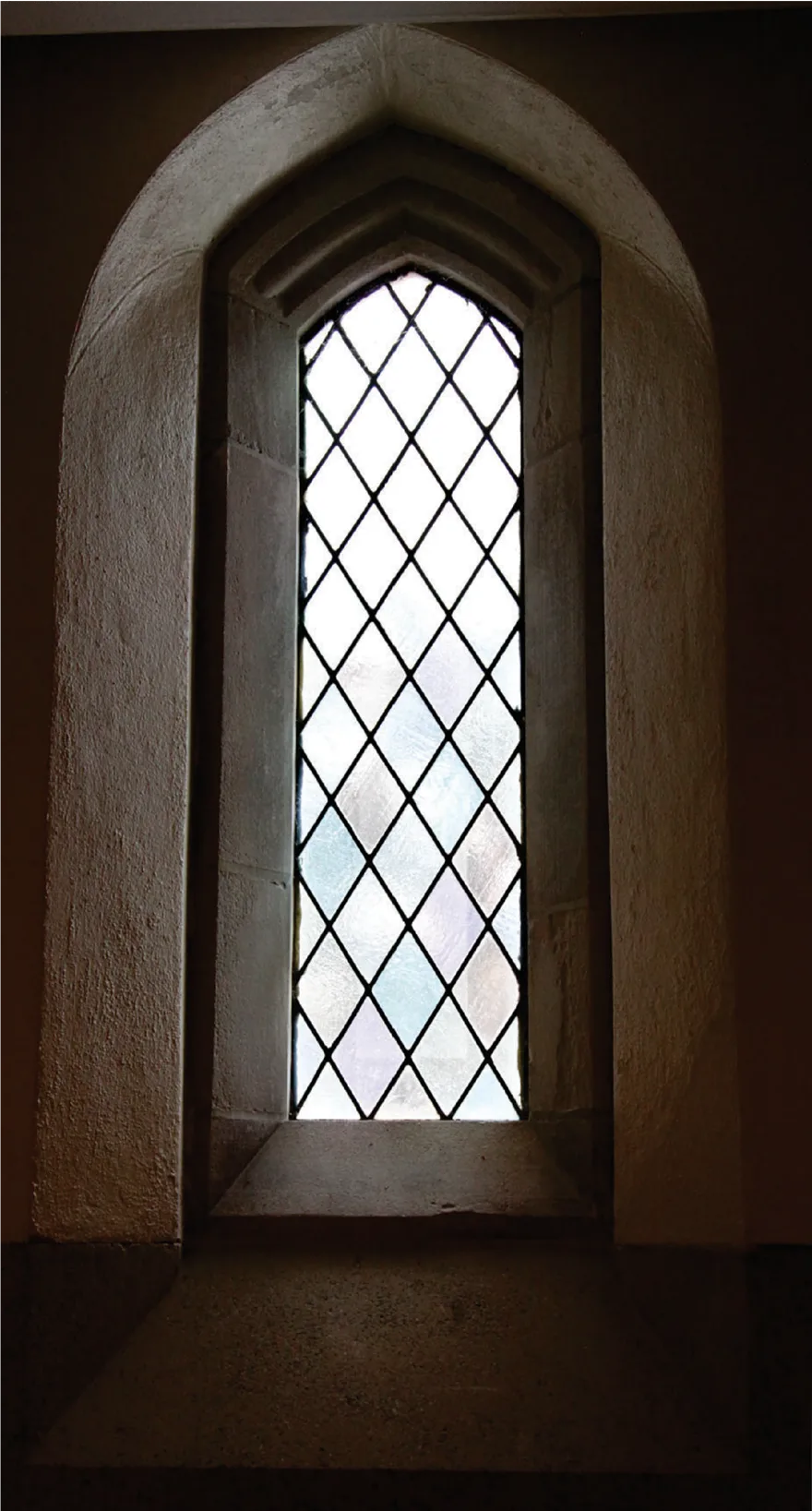 Tall pointed-arch window of diamond-paned leaded glass set deep in a pale stone wall, glowing against the dark interior
